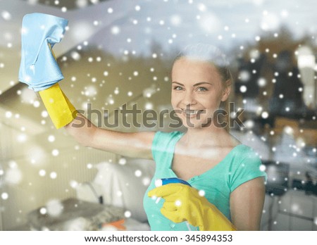 people, housework and housekeeping concept - happy woman in gloves cleaning window with rag and cleanser spray at home over snow effect