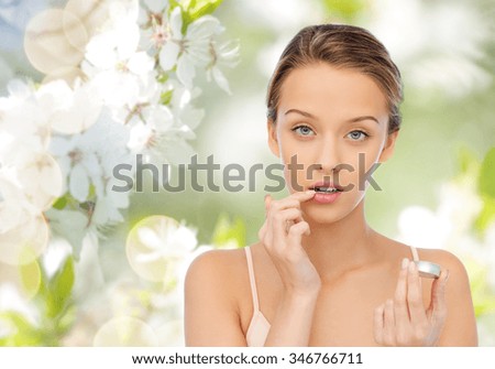 beauty, people and lip care concept - young woman applying lip balm to her lips over green natural background with cherry blossoms