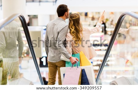 sale, consumerism and people concept - happy young couple with shopping bags going down by escalator and pointing finger in mall
