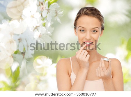 beauty, people and lip care concept - smiling young woman applying lip balm to her lips over green natural cherry blossom background