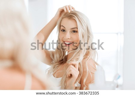 beauty, hygiene, hairstyle, morning and people concept - smiling young woman looking to mirror at home bathroom