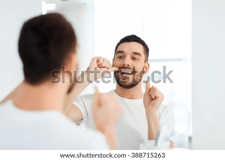 health care, dental hygiene, people and beauty concept - smiling young man with floss cleaning teeth and looking to mirror at home bathroom