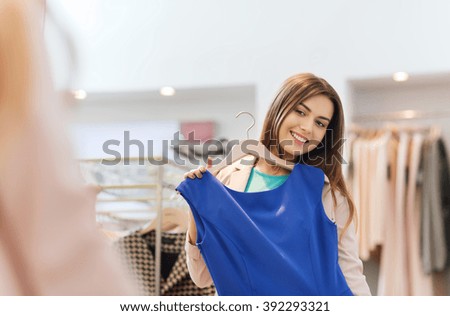 shopping, fashion, style and people concept - happy woman choosing clothes and looking to mirror in mall or clothing store