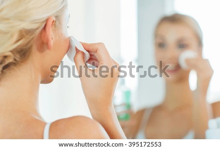 beauty, skin care and people concept - close up of smiling young woman cleaning her face with cotton disc and lotion at bathroom