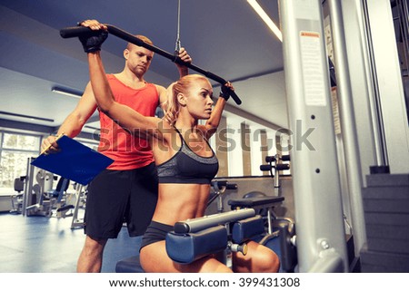 sport, fitness, teamwork and people concept - young woman flexing muscles on gym machine and personal trainer with clipboard
