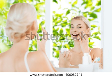beauty, skin care and people concept - smiling young woman applying cream to face and looking to mirror at home bathroom over green natural background