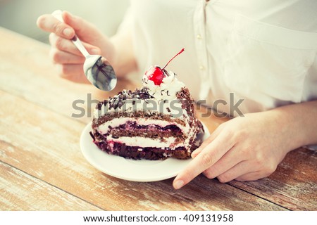 food, junk-food, culinary, baking and holidays concept - close up of woman eating chocolate cherry cake with spoon and sitting at wooden table