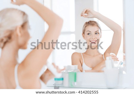 beauty, hygiene, morning and people concept - smiling young woman applying antiperspirant or stick deodorant and looking to mirror at home bathroom