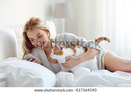 technology, pets, communication and people concept - happy young woman with cat and smartphone texting message in bed at home