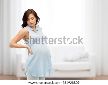 pregnancy, health, people and expectation concept - pregnant woman touching her back and suffering from backache over home living room background