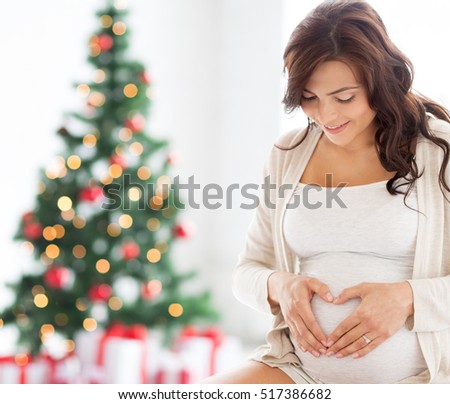 pregnancy, love, holidays, people and expectation concept - happy pregnant woman making heart gesture at home over christmas tree background