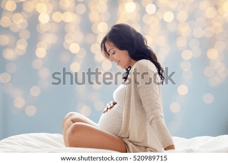pregnancy, rest, people and expectation concept - happy pregnant woman sitting on bed and touching her belly over holidays lights background