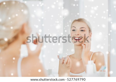beauty, skin care and people concept - smiling young woman applying cream to face and looking to mirror at home bathroom over snow