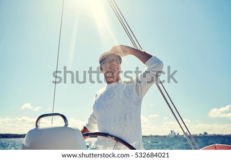 sailing, age, tourism, travel and people concept - happy senior man in captain hat on steering wheel and looking far away sail boat or yacht floating in sea