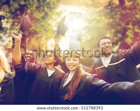 education, graduation and people concept - group of happy international students in bachelor gowns waving mortar boards or hats over campus background