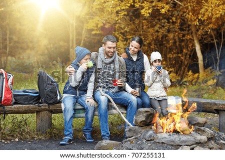 camping, travel, tourism and people concept - happy family sitting on bench and drinking hot tea from cups at camp fire in autumn forest