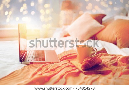 technology, holidays, christmas, interior and winter concept - cozy bedroom with laptop computer, coffee cup and croissant on bed at cozy home