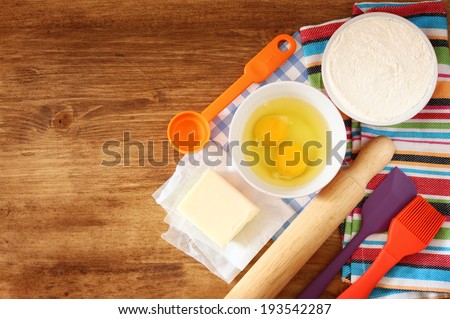 top view of baking ingredients - dough, eggs, flour, butter, and kitchen utensils  over wood board.