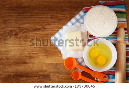 top view of baking ingredients - dough, eggs, flour, butter, and kitchen utensils over wood board.
