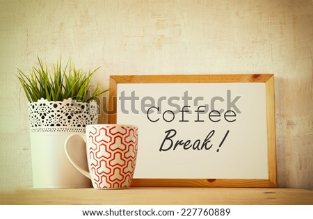 white drawing board with the phrase coffee break over wooden table with coffe cup and flower pot decoration . filtered image