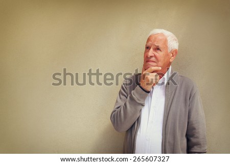 Thoughtful senior man looking up into the air  as he stands against a blank textured wall  with copyspace