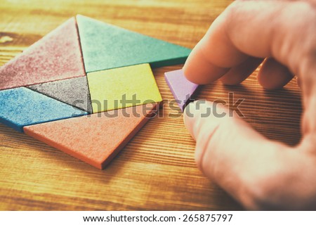 man\'s hand holding a missing piece in a square tangram puzzle, over wooden table.