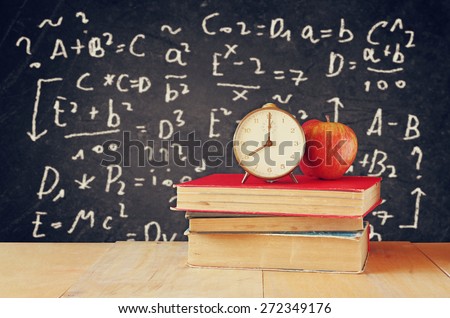 image of school books on wooden desk, apple and vintage clock over black background with formulas. education concept