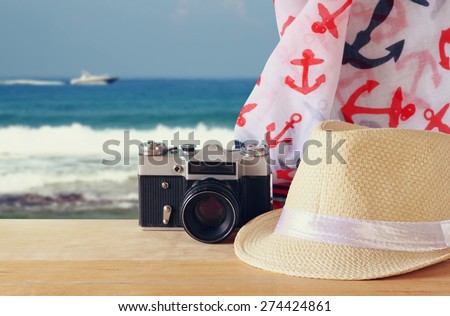 fedora hat, old vintage camera and scarf over wooden table and sea landscape background. relaxation or vacation concept