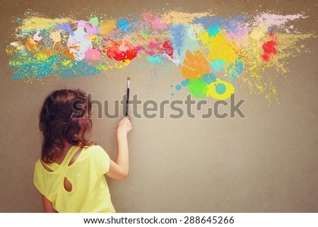 back view cute kid holding brush next to textured wall and paint splashes