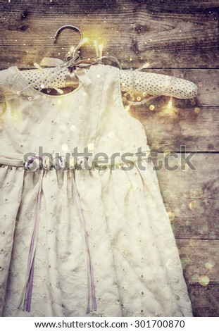 abstract image of vintage cream girl\'s dress on hanger with on wooden background with garland lights. glitter overlay