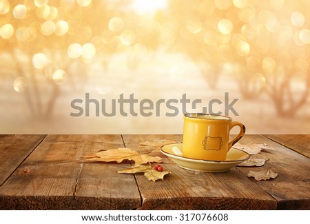 front image of coffee cup over wooden table and autumn leaves in front of autumnal sunset background