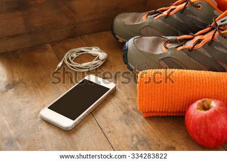 fitness concept with mobile phone with earphones, towel, apple and sport footwear over wooden background.