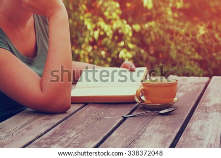 close up image of woman reading book outdoors, next to wooden table and coffe cup at afternoon. filtered image. filtered image. selective focus