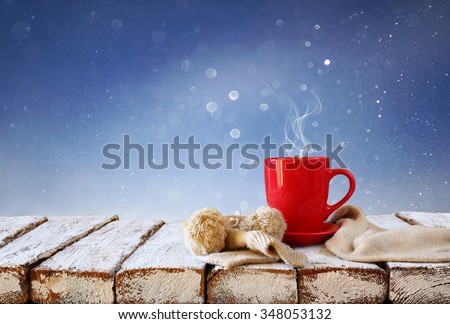 Cup of hot coffee and cozy knitted scarf on wooden table in front of glitter background