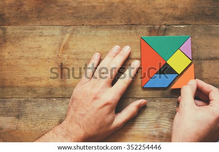 top view photo of man\'s hand holding a missing piece in a square tangram puzzle, over wooden table.