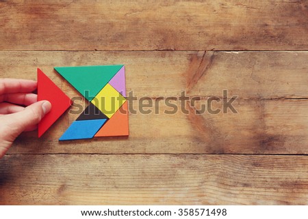 man\'s hand holding a missing piece in a square tangram puzzle, over wooden table.