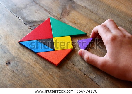 kid\'s hand holding a missing piece in a square tangram puzzle, over wooden table.