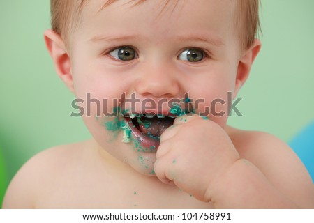 green eyed boy smiling with his hand in his mouth, with his hand and face covered in green icing and cake. with a blue balloon in the background on a green backdrop.