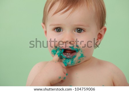 green eyed boy smiling with his finger in his mouth, with hand and face covered in green icing and cake on a green backdrop.