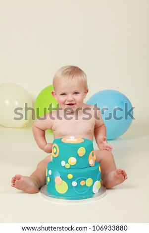 Blond hair blue eyed baby boy being cheeky smiling and pulling silly faces by his blue yellow green and orange two tier birthday party cake while sitting on a cream background with balloons behind