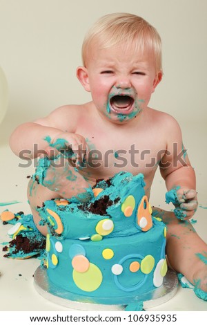 Blond hair blue eyed baby boy crying and screaming while eating his blue yellow green and orange two tear butter birthday party cake while sitting on cream background with foot in mess smearing icing