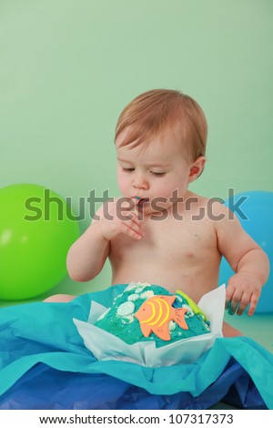 Cute happy brunette baby boy with closed eyes is tasting his blue butter icing on his ocean themed giant fish birthday cupcake licking sticky icing off his fingers and sitting on green background
