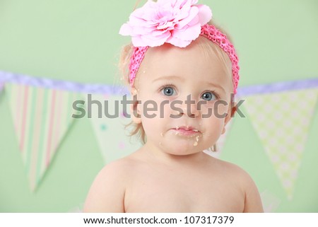 Adorable blond hair blue eyed baby girl with pink flower head band eating and tasting vanilla sponge cake while sitting on green background with pink green white purple and blue flag bunting behind