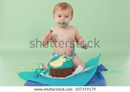 Adorable and surprised smiling brunette haired baby boy with blue butter icing on face holding cake with his sticky fingers and his ocean themed giant cupcake broken up on seamless green background
