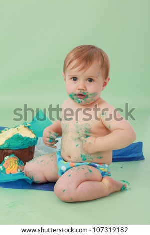 Cute and cuddly brunette haired baby boy with face covered in blue butter icing touching his sticky tummy with hand and wearing polka dot blue pants with giant cupcake in the background all smashed up