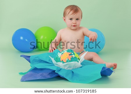 Brunette hair blue eyed baby boy holding hand up and sitting in front of his blue yellow green and orange giant fishy birthday cupcake with tissue paper on a seamless green background with balloons