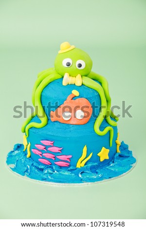 Sea themed birthday party cake with green octopus with hat bow tie and eyes and orange submarine and pink fish on two round blue double tier butter iced cake isolated on a green seamless background