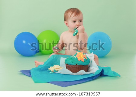 Brunette hair blue eyed baby boy eating and tasting the icing of his ocean themed birthday cupcake with orange fish and seaweed while sitting on green background with green and blue balloons behind