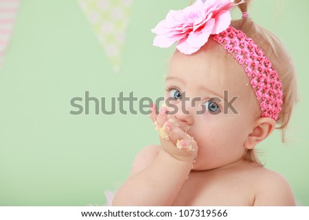 Adorable blond hair big blue eyed baby girl with pink flower elastic head band on head and pony tail in hair sucking eating and tasting vanilla sponge cake on green background with flag bunting behind
