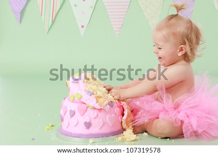 Adorable laughing blond haired baby girl with pony tail in pink tutu touching vanilla sponge cake with pink and purple fondant heart icing while sitting on green background with flag bunting behind